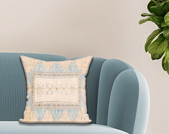 Puede incluir: Un cojín cuadrado decorativo con un patrón geométrico beige y azul claro. El cojín está colocado en un sofá azul verdoso. El diseño del cojín presenta un motivo rectangular central con detalles y bordes.