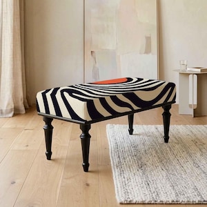 May include: A rectangular ottoman featuring a bold black and cream-colored swirling pattern across its top surface, accented by a single solid orange semi-circle. The piece is supported by four slender, tapered black wooden legs.