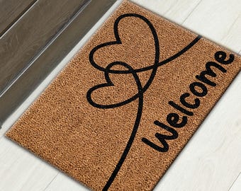 Puede incluir: Una alfombra de coco marrón con la palabra "Welcome" y dos corazones entrelazados en negro.