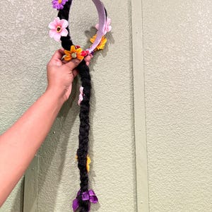 May include: A black braided hair accessory with a floral headband. The headband features pink, purple, and yellow flowers, and a silver crown. The braid is adorned with additional flowers and purple ribbons at the end.