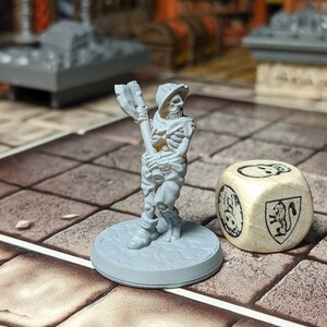 May include: A gray plastic miniature skeleton figure holding a weapon, standing on a gray base. A wooden dice with a skull and shield design is in the background.