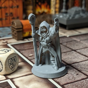 May include: A gray plastic miniature of a skeletal wizard holding a staff, wearing a robe and standing on a gray base. The miniature is on a brown and white tiled surface with a white wooden dice with black markings.