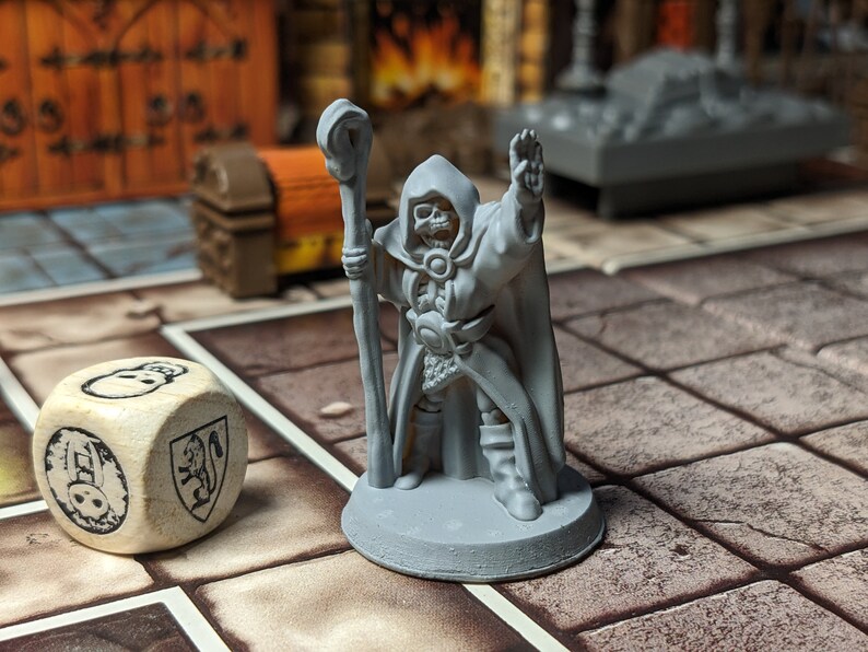 May include: A gray plastic miniature of a skeletal wizard holding a staff, wearing a robe and standing on a gray base. The miniature is on a brown and white tiled surface with a white wooden dice with black markings.
