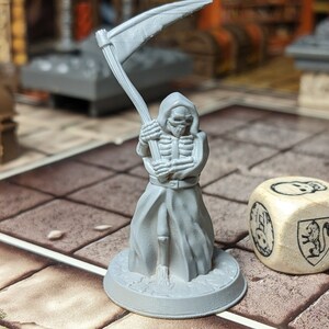 May include: A gray plastic miniature of a skeletal figure in a hooded robe, holding a scythe. The figure is standing on a round base. A wooden dice with a skull and a lion symbol is in the foreground.