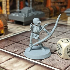 May include: A gray plastic miniature skeleton archer figure holding a bow and arrow, standing on a gray base. The figure is posed as if it is aiming the bow and arrow. A white wooden dice with a skull and a squirrel design is in the background.
