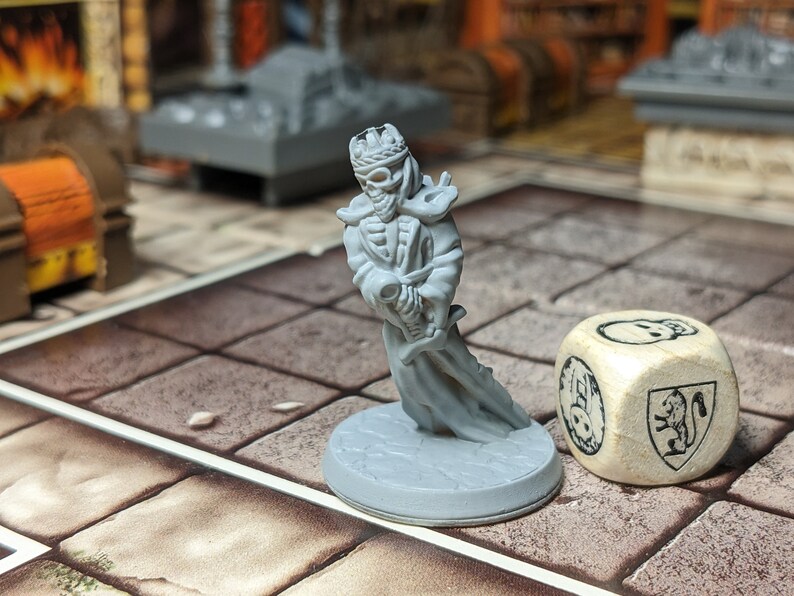 May include: A gray plastic miniature of a skeletal figure wearing a crown and holding a sword. The figure is standing on a gray base. A wooden dice with a skull and a coat of arms is in the background.