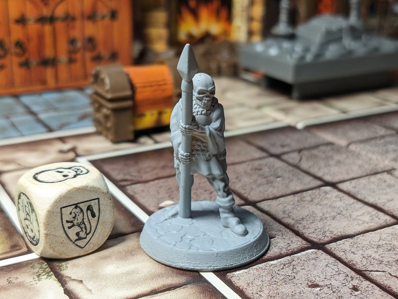 May include: A gray plastic miniature figure of a skeletal warrior holding a spear. The figure is standing on a round base. A wooden dice with a skull and a lion crest is in the foreground.