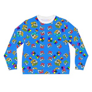 Op de afbeelding: Een lange mouwen sweatshirt met een helderblauwe achtergrond, bedekt met een herhalend patroon van kleurrijke Rubik's Cubes. De manchetten en de halslijn zijn wit, wat een contrasterende rand creëert. Het ontwerp toont verschillende kubusoriëntaties en kleurencombinaties.