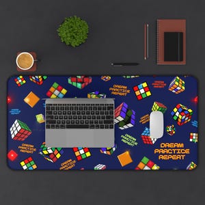 Op de afbeelding: Een blauwe bureaumat met een patroon van kleurrijke Rubik's Cubes en de tekst "DREAM PRACTICE REPEAT" in oranje. Een laptop, muis en andere kantoorbenodigdheden liggen op het donkere oppervlak.