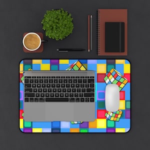 Könnte beinhalten: Ein Laptop, eine Maus und ein Mauspad im Rubik's Cube-Design. Das Mauspad hat ein lebendiges Muster aus Quadraten und Rubik's Cubes. Eine Tasse Kaffee, ein Notizbuch und ein Stift befinden sich ebenfalls auf der dunklen Oberfläche.