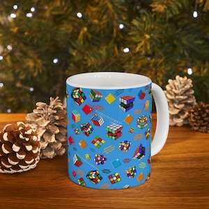 Puede incluir: Taza de cerámica blanca con un fondo azul cubierto de coloridos diseños de Cubo de Rubik. La taza tiene un asa blanca y un interior blanco. La taza está sobre una superficie de madera con piñas y vegetación en el fondo.