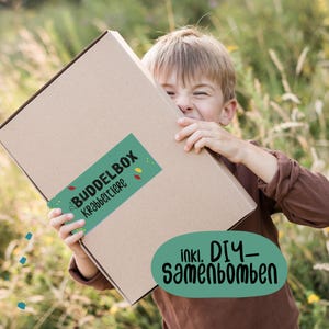 Könnte beinhalten: Ein Kind hält einen großen, rechteckigen Karton. Auf dem Karton befindet sich ein grüner Aufkleber mit der Aufschrift "BUDDELBOX Krabbeltiere" und ein grüner ovaler Aufkleber mit der Aufschrift "inkl. DIY-Samenbomben". Der Hintergrund ist verschwommen mit grünen und gelben Farbtönen.