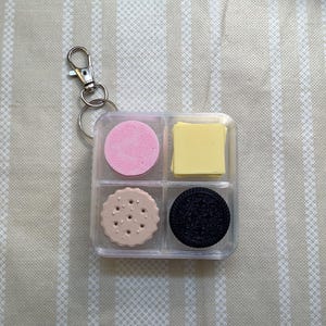 May include: A clear square plastic container with a metal keychain attachment, divided into four compartments. Inside are four distinct items: a pink circular disc, a stack of yellow square sheets, a beige scalloped cookie, and a black sandwich cookie.