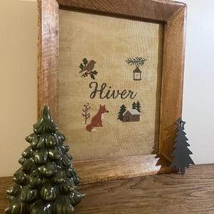 Peut inclure: Un cadre avec une broderie au point de croix avec le mot "Hiver" et des images sur le thème de l'hiver. Un sapin de Noël vert en céramique et un arbre en métal noir sont devant le cadre. Le cadre est en bois brun clair.