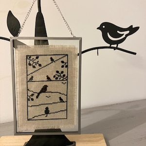 Peut inclure: Une broderie au point de croix encadrée représentant une série d'oiseaux noirs sur fond blanc. Le cadre est argenté et possède un pied noir. La pièce est suspendue à une branche en métal noir avec une silhouette d'oiseau noir.