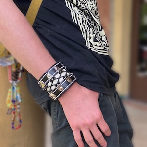 May include: A black leather bracelet with white stitching and silver studs. The bracelet has a criss-cross design and is adjustable.