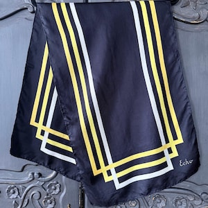 Puede incluir: Una bufanda negra con un diseño geométrico de rayas y cuadrados amarillos y blancos. La bufanda está colgada de una percha negra. La palabra "Echo" está impresa en la bufanda.