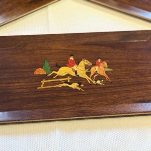 May include: A rectangular wooden tray with a dark brown finish. The tray features an inlaid scene of two riders on horseback, a dog, and a fence. The riders wear red coats. The horses are yellow and brown.