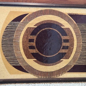 May include: Abstract geometric artwork in a dark wooden frame. The artwork features concentric circles and shapes in shades of brown, beige, and dark burgundy. The design incorporates textured materials, creating a layered effect. The artist's signature is visible in the upper right corner.