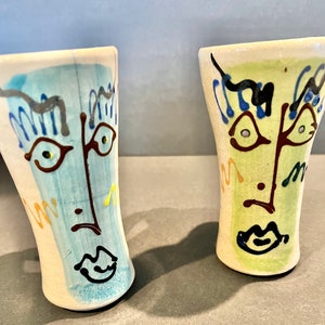 May include: Two ceramic cups with abstract faces painted on them. One cup is blue and white with a yellow accent, the other is green and white with a brown accent.