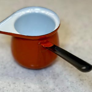 May include: A small, orange enamelware saucepan with a black handle. The interior is light blue, and the rim is white. The spout is designed for easy pouring. This vintage-style pot is ideal for heating liquids.