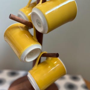Puede incluir: Cuatro tazas de cerámica amarillas con interiores blancos cuelgan de un árbol de tazas de madera. Las tazas tienen un acabado brillante y asas curvas. El árbol de tazas es de madera oscura y tiene una base redonda.