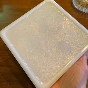May include: White square glass dish with a raised floral design. The dish has a textured surface and a slightly iridescent finish.