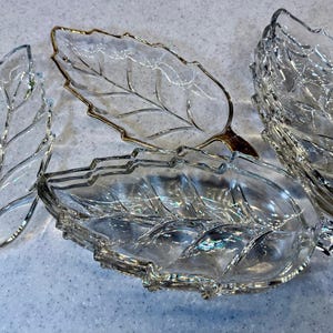 Puede incluir: Cuatro fuentes de vidrio transparente con forma de hoja. Dos tienen ribetes plateados, una tiene ribetes dorados y otra no tiene ribetes. Los platos están dispuestos sobre una superficie clara, mostrando los detalles de las venas de las hojas.