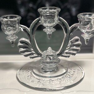 May include: A clear glass three-arm candelabra with a decorative base and ornate details.