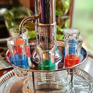 Könnte beinhalten: Ein Vintage-Likörspender aus Chrom und Glas mit einem runden Tablett, das Schnapsgläser enthält. Die Schnapsgläser sind mit farbigen Bändern in Rot, Blau und Grün verziert. Der Spender hat einen hohen, polierten Metallkörper und einen Auslauf.