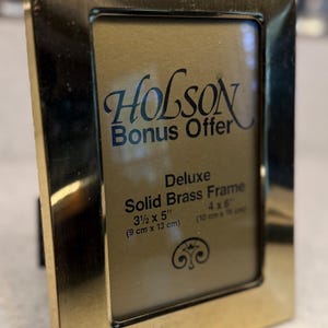 May include: A rectangular brass photo frame with the text "HOLSON Bonus Offer" and "Deluxe Solid Brass Frame". The frame is 3 1/2 x 5 inches (9 cm x 13 cm) and is designed to hold a 4 x 6 inch (10 cm x 15 cm) photo.