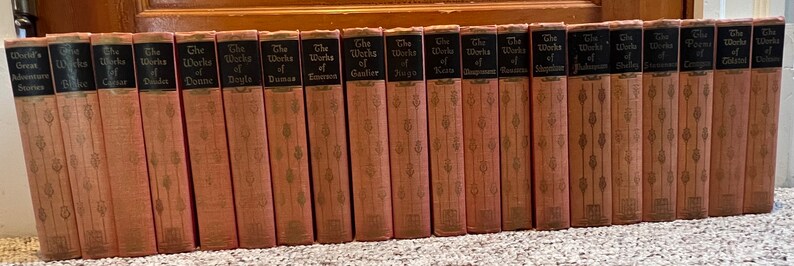 May include: A set of 20 vintage hardcover books with spine titles including "The Works of" followed by the author's name. The books are all the same size and have a light pink cover with a gold design.