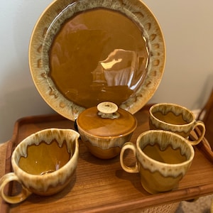 May include: A set of brown and white glazed ceramic teacups, a creamer, and a sugar bowl with a lid. The set is on a wooden tray.