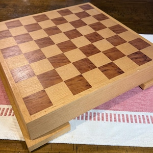 May include: Wooden chess board with a checkerboard pattern of light and dark brown squares. The board is made of wood and has a rectangular shape. The chess board is sitting on a red and white striped surface.