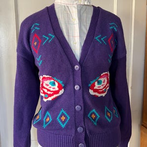 May include: Purple cardigan sweater with a geometric pattern and floral accents. The cardigan has a V-neck and button closure.