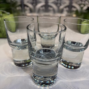 Puede incluir: Primer plano de cinco vasos de chupito de cristal transparente. Los vasos tienen una base gruesa y un borde ligeramente ensanchado. Los vasos de chupito están vacíos y parecen limpios. El fondo está borroso, con un diseño de azulejos.