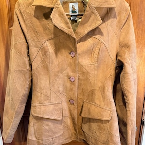 May include: A tan suede blazer with a patchwork design. The jacket has a notched collar, two front pockets, and three buttons. The blazer is hanging on a wooden door.