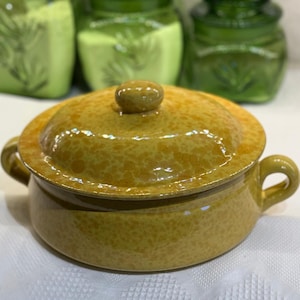 May include: A yellow ceramic soup bowl with a lid and two handles. The bowl has a speckled pattern and a small knob on the lid. Other green ceramic jars are visible in the background.