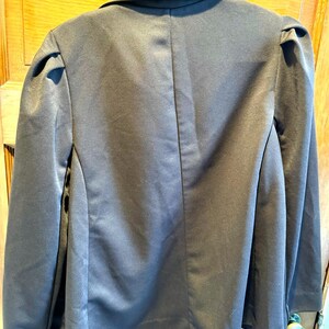 Puede incluir: Una blazer marrón oscuro con solapas de muesca y hombros acolchados cuelga de una percha negra. La blazer tiene un brillo satinado y un corte entallado, lo que sugiere un estilo formal o semi-formal.