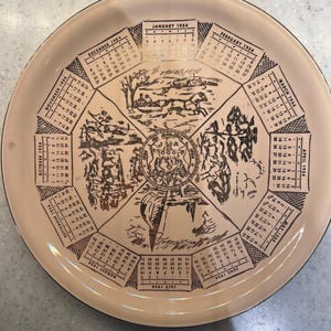 May include: A round ceramic plate with a beige background and gold trim. The plate features a calendar for each month of 1954, with illustrations of scenes from each month. The plate is decorated with a gold border and a gold seal in the center.