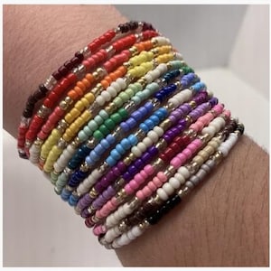 May include: A selection of seed bead bracelets in a rainbow of colours, stacked on a wrist. The bracelets display shades of red, orange, yellow, green, blue, purple, pink, white, and black. Each bracelet is made of small, round beads.