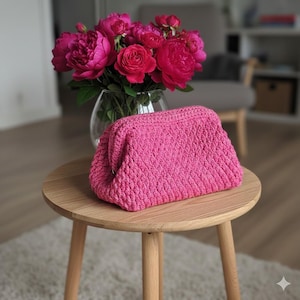 Peut inclure: Une pochette rose vif, crochetée à la main, avec un motif texturé, posée sur une petite table en bois. Le sac est rectangulaire avec des bords arrondis. Un vase de pivoines roses vives est en arrière-plan.