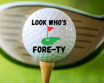 May include: A white golf ball with the text "Look Who's Fore-Ty" printed on it. The ball is on a tee and a golf club is behind it.