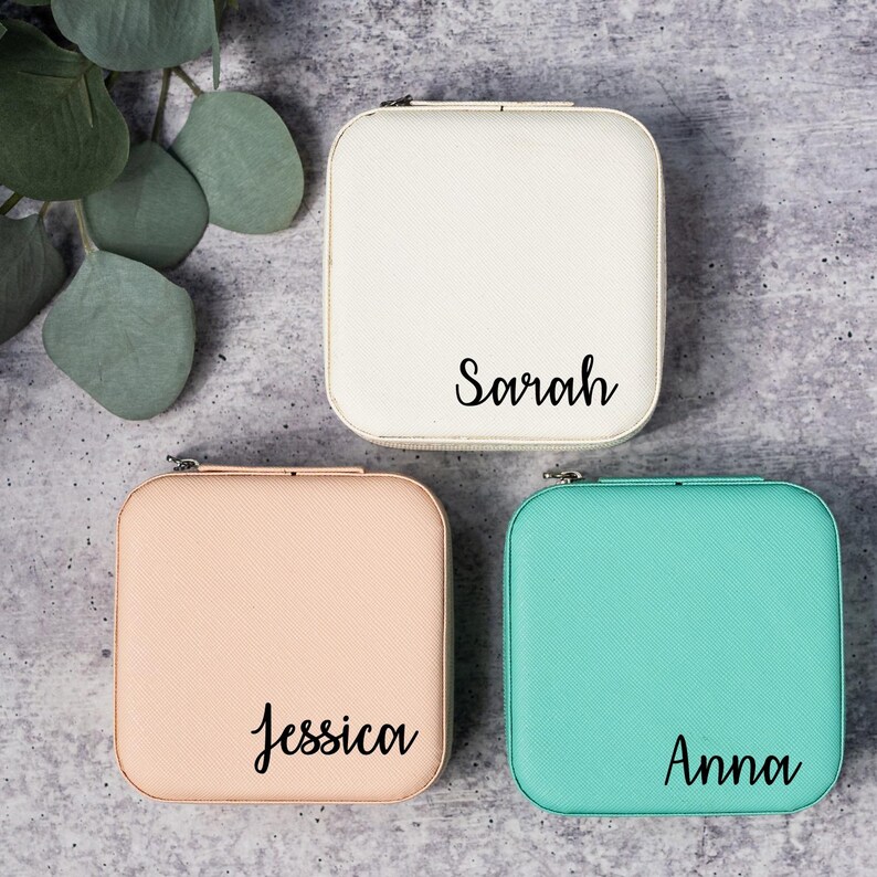 May include: Three square jewelry boxes with personalized names. The boxes are white, light pink, and light blue.