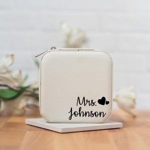 May include: A white jewelry box with a zipper closure. The box has a black inscription that reads "Mrs. Johnson" with two black hearts.