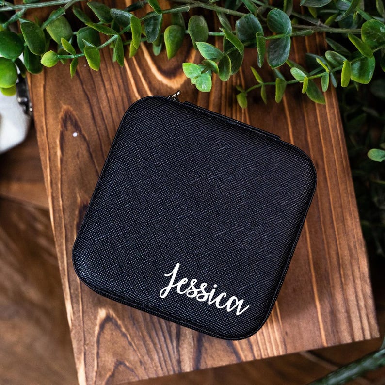 May include: A black square-shaped jewelry box with a zipper closure. The box is made of a textured material and has the name "Jessica" printed in white on the front.