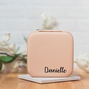 May include: A light pink square-shaped jewelry box with a zipper closure. The box has a textured surface and the name "Danielle" is printed in black on the front.