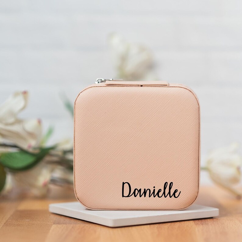 May include: A light pink square-shaped jewelry box with a zipper closure. The box has a textured surface and the name "Danielle" is printed in black on the front.
