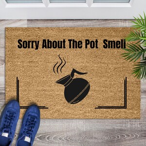 May include: A rectangular doormat with the text "Sorry About The Pot Smell" above a black coffee pot illustration. The mat is brown with a textured surface and black corner accents. Blue sneakers are visible at the bottom.