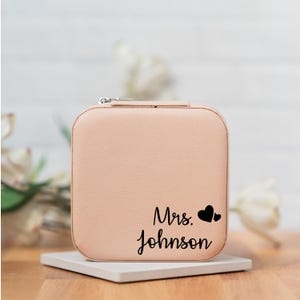 May include: A light pink jewelry box with a zipper closure. The box has the text "Mrs. Johnson" and two small hearts printed on the front.